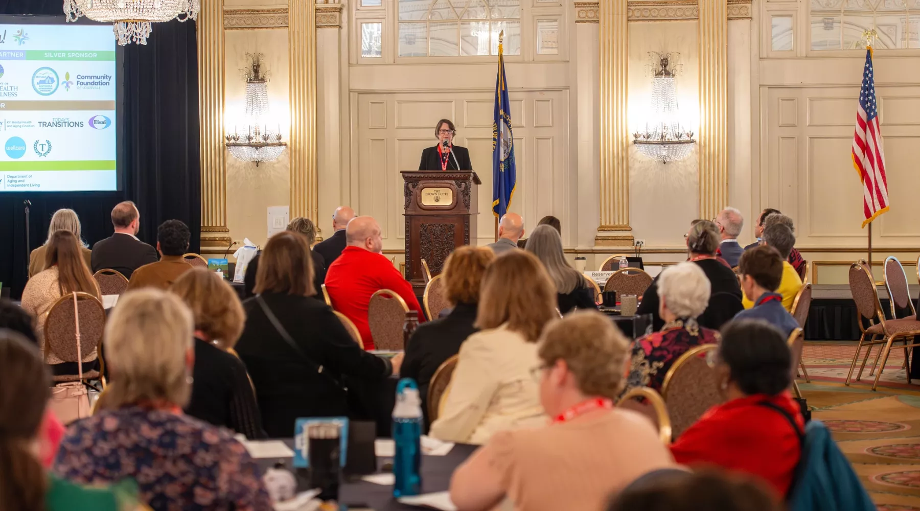 View of a conference room full of attendees seated and listening to a female speaker