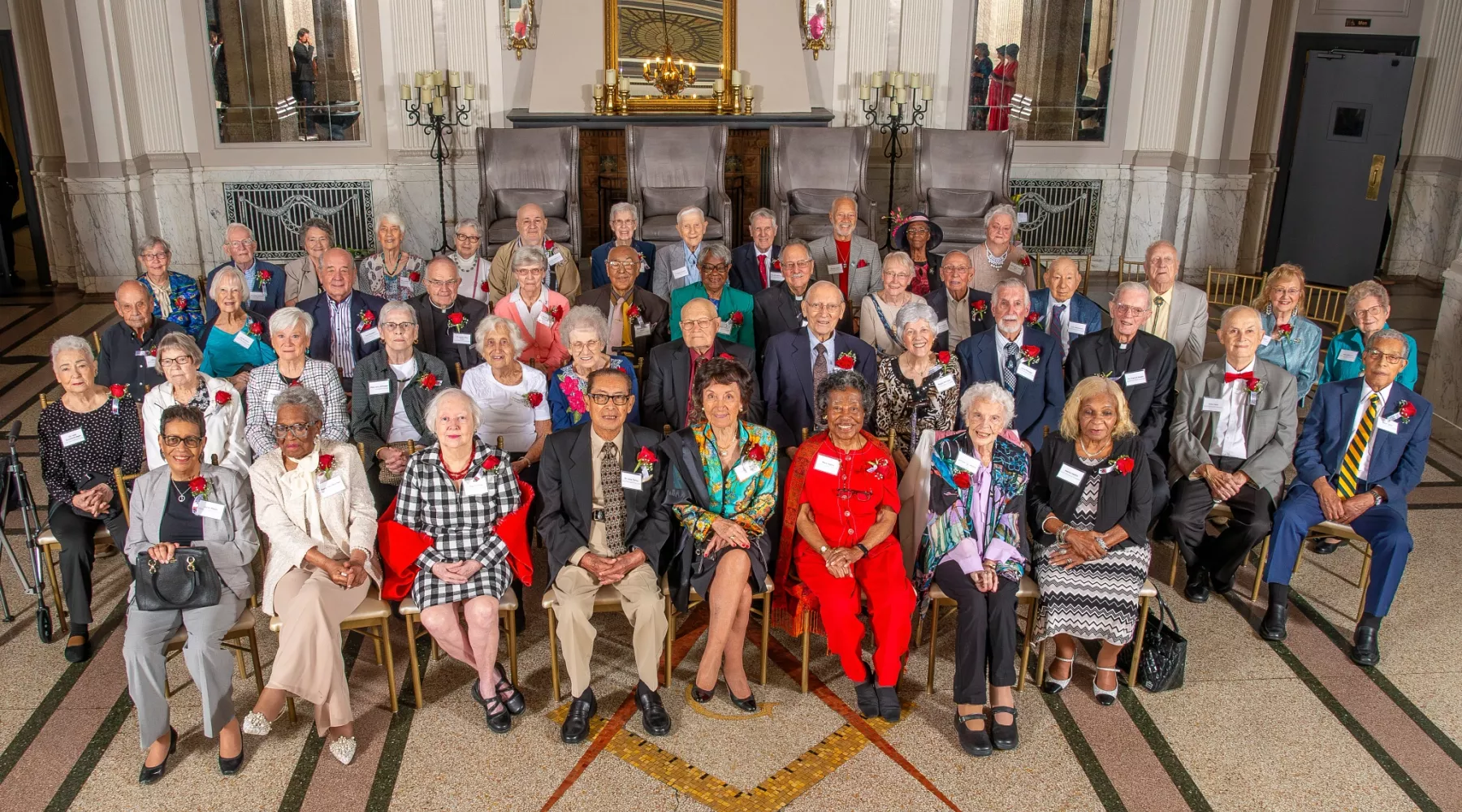 A group of older adults wearing red rose boutonnieres assembled in chairs and smiling for a photo