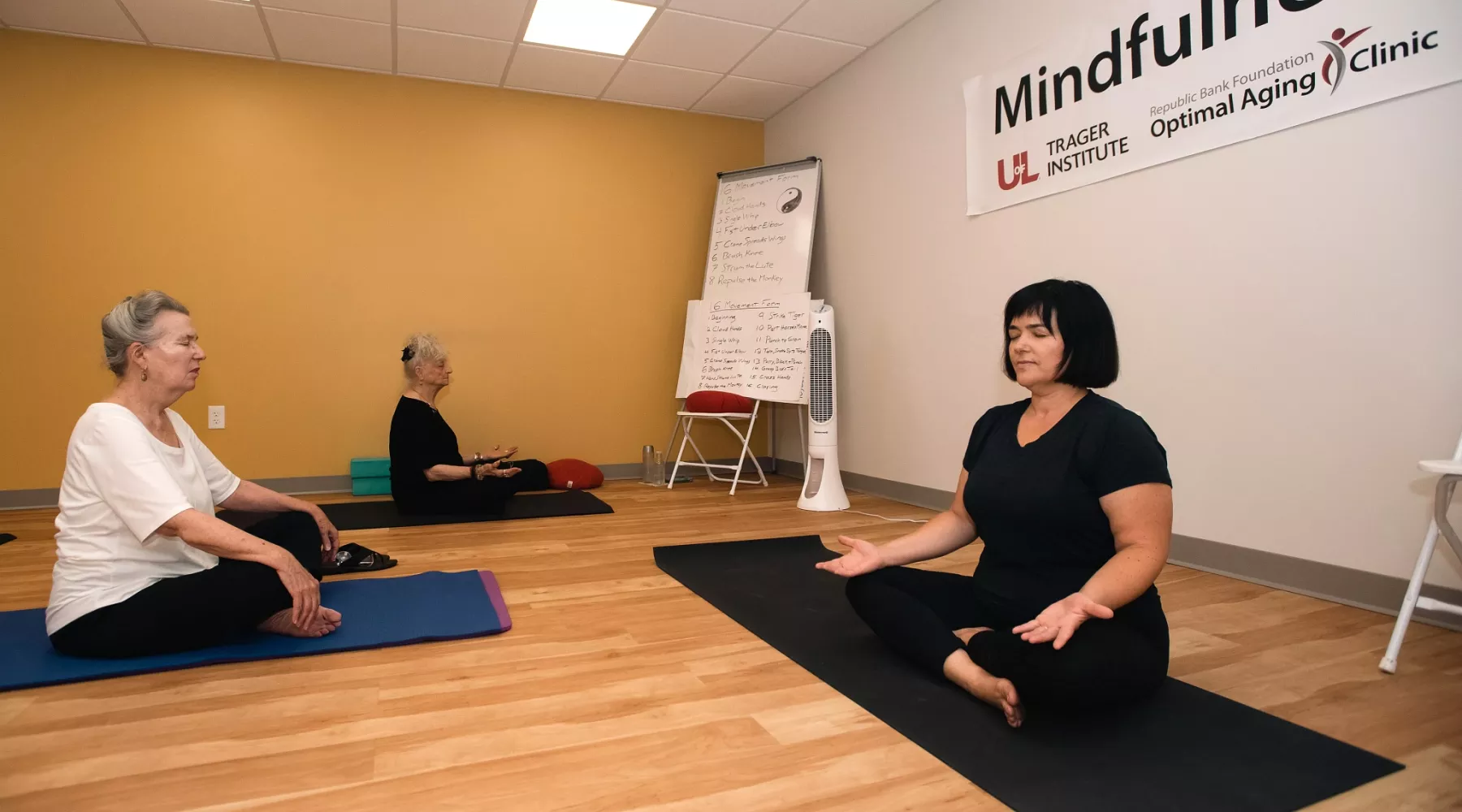 Two older adult women sitting in front of a younger woman yoga instructor