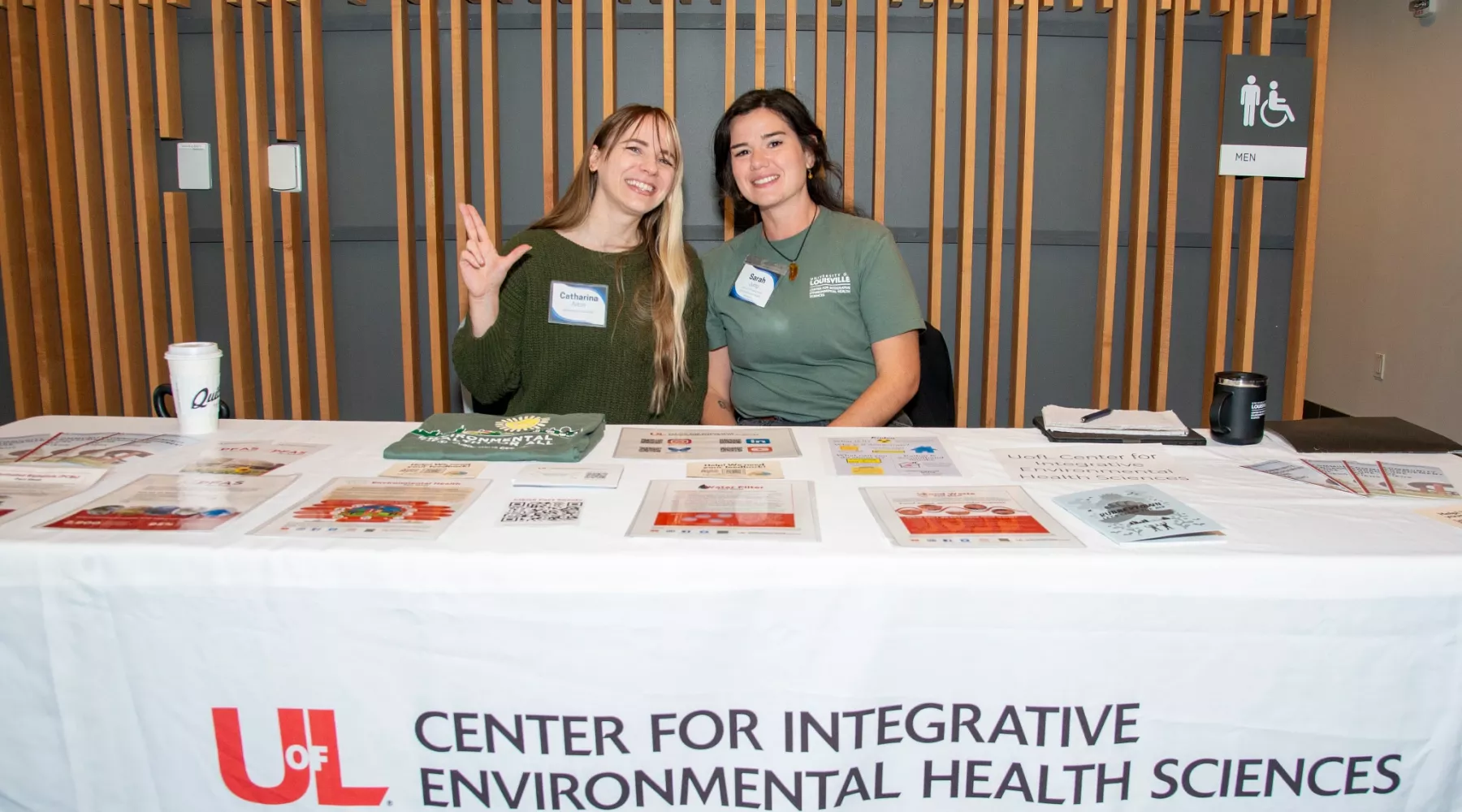 Two people smile while seated behind an outreach table for the University of Louisville Center for Integrative Environmental…