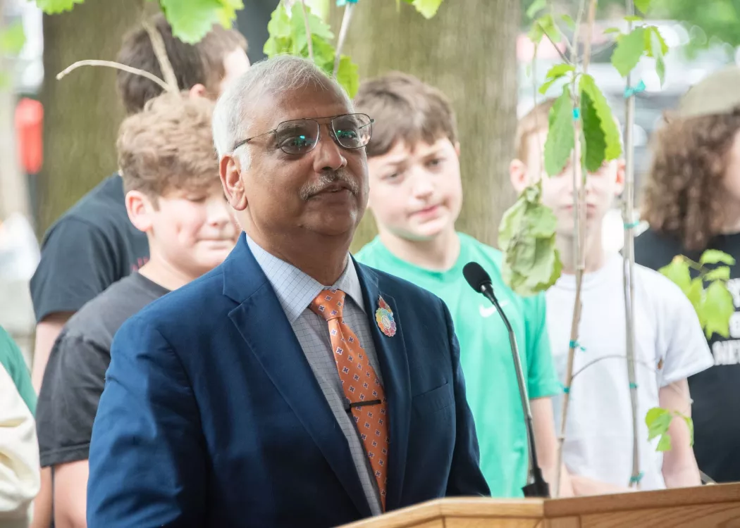 Man speaking at a podium with children and trees in background.