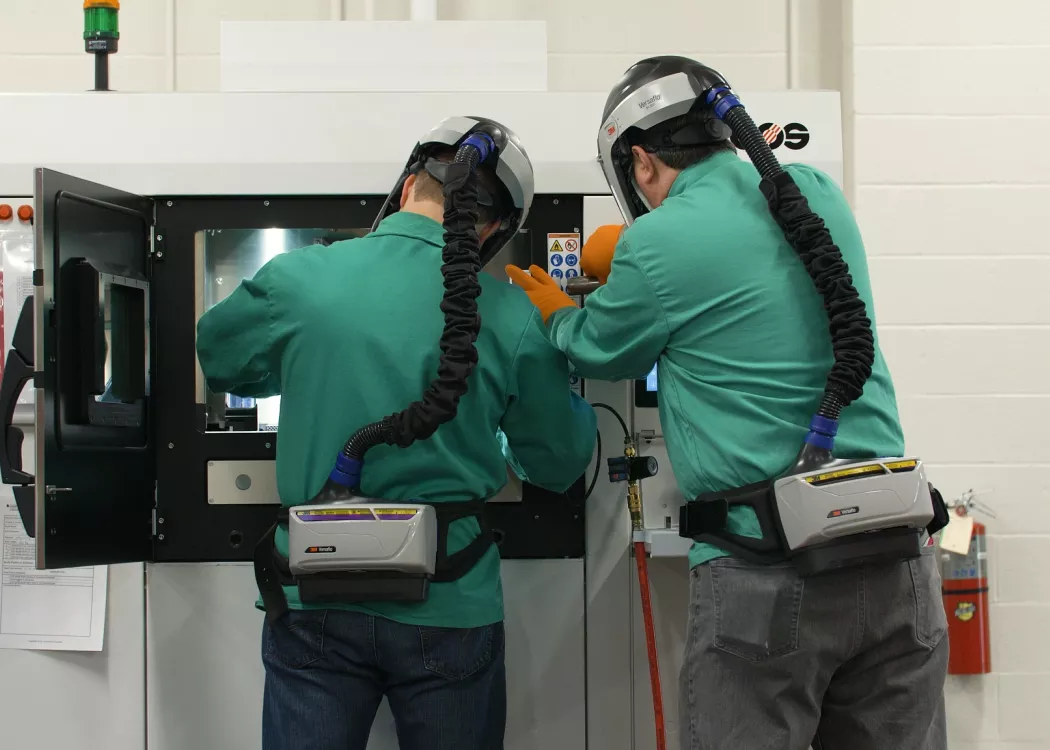 Two trainees wearing respirator helments while working with additive manufacturing machinery at AMIST.